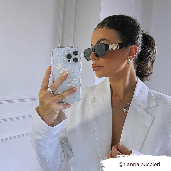A woman with a sleek ponytail takes a mirror selfie while wearing black sunglasses, a white blazer, and minimal jewelry. She holds an iPhone15 with the Coconut Lane Pearls & Bows Phone Case, decorated with white floral designs. The Instagram handle "@tianna.buccieri" is displayed in the bottom right corner.