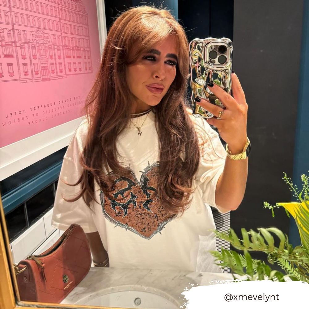 A person with long brown hair takes a mirror selfie in a stylish indoor setting. They wear a white shirt with a detailed design and hold their phone, which is adorned with Coconut Lane's 3D Chrome Phone Case. A pink handbag and some greenery are visible in the foreground. Text in the image reads "@xmevelynt.
