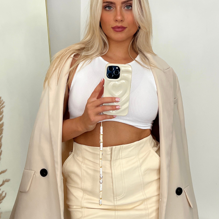 A woman with long blonde hair holds up her phone, taking a mirror selfie. She is wearing a white cropped tank top, a light beige skirt, and a matching beige coat draped over her shoulders. Her phone sports the stylish Melting Heart Phone Case - Coconut Cream by Coconut Lane. The background is plain white, with a dry plant partially visible on the left.