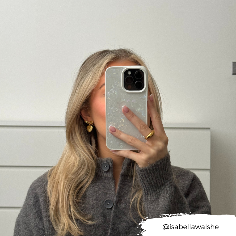 A person with long, blonde hair is taking a mirror selfie with their smartphone, which is adorned with the light-colored Mother of Pearl Phone Case by Coconut Lane. They are wearing a grey sweater and gold hoop earrings. The Instagram handle "@isabellawalshe" is displayed at the bottom right.