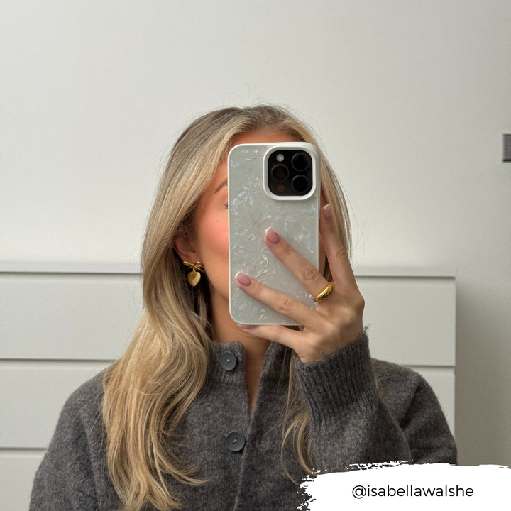 A person with long, blonde hair is taking a mirror selfie with their smartphone, which is adorned with the light-colored Mother of Pearl Phone Case by Coconut Lane. They are wearing a grey sweater and gold hoop earrings. The Instagram handle "@isabellawalshe" is displayed at the bottom right.