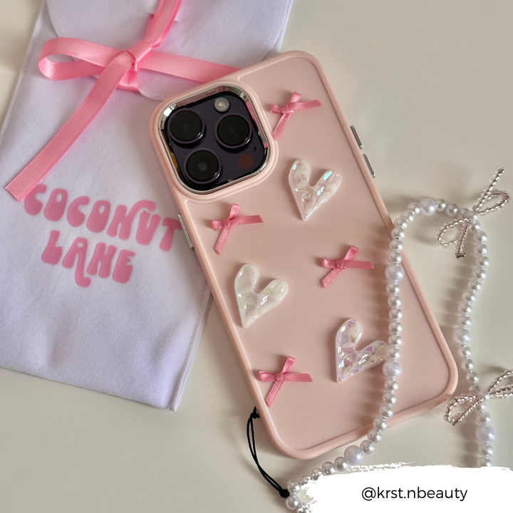 The Pearly Hearts Phone Case - Pink from Coconut Lane is a protective accessory for your iPhone 15, adorned with decorative pearl hearts and pink ribbons. It is displayed next to a pink pouch labeled "COCONUT LANE," with a pearl necklace featuring a bow nearby. The Instagram handle @krst.nbeauty is visible in the bottom right corner.