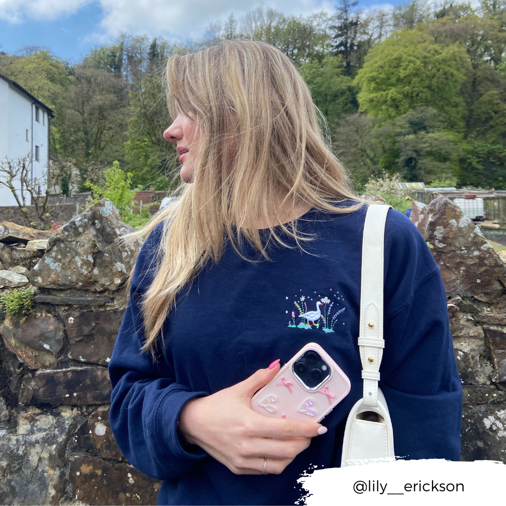 A person with long hair wearing a navy blue sweatshirt holds an iPhone15 encased in Coconut Lane's Pearly Hearts Phone Case - Pink and stands outdoors beside a stone wall. They have a white bag over their shoulder and are looking to the left, with trees and a building visible in the background. Instagram handle @lily__erickson is on the bottom right.