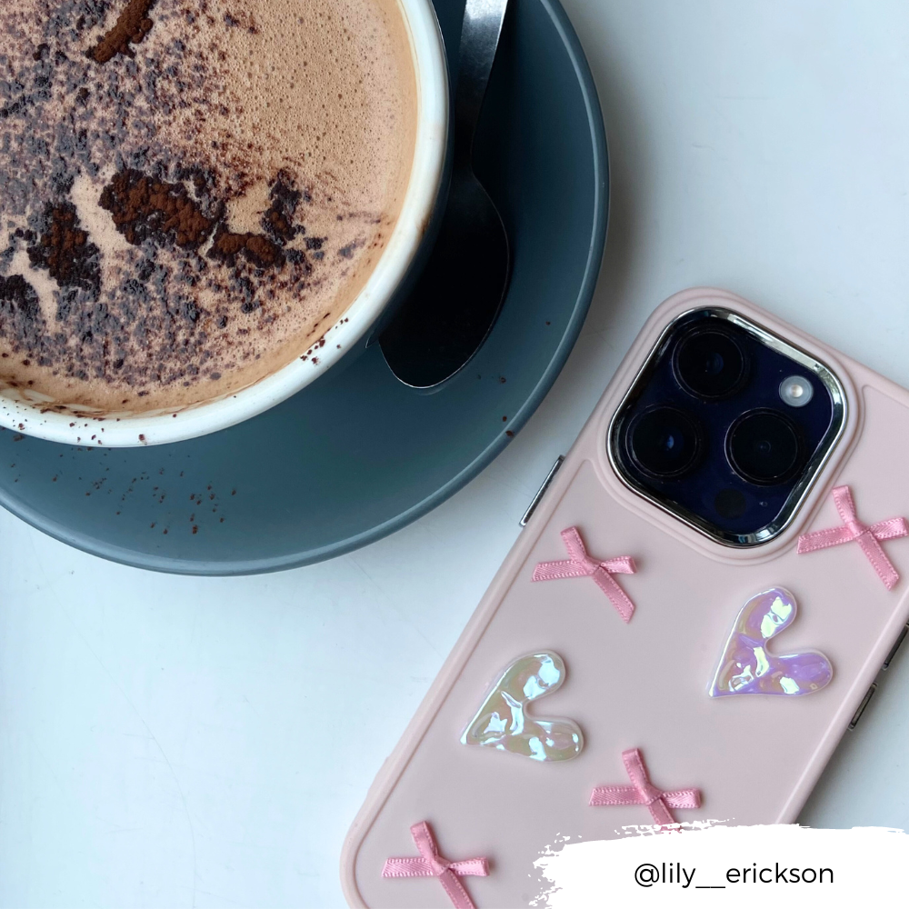 A cup of frothy hot chocolate in a white mug sits on a grey saucer with a spoon. Next to it is the Pearly Hearts Phone Case - Pink from Coconut Lane, decorated with pink bows and shiny heart stickers. The Instagram handle "@lily__erickson" appears at the bottom of the image.