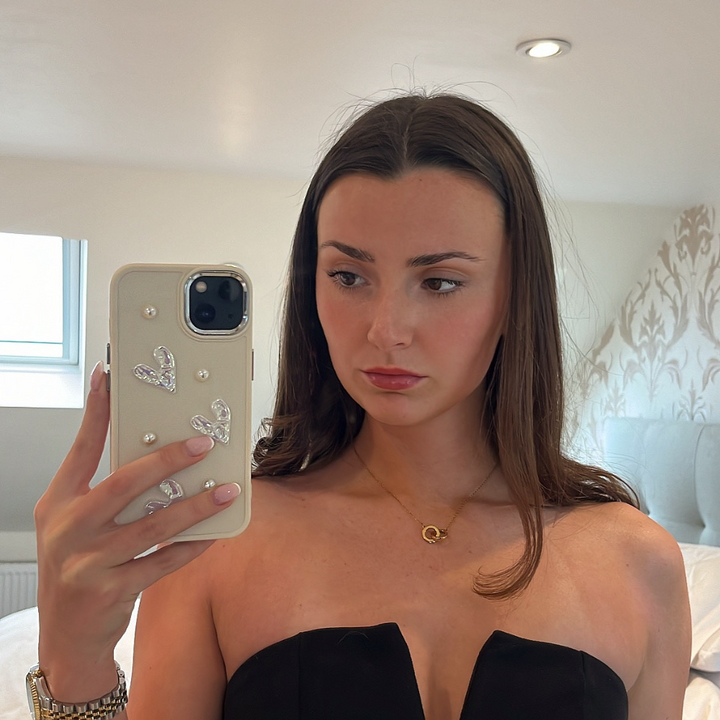 A woman with long brown hair, wearing a strapless black top and a gold necklace, is taking a selfie in a mirror. She is holding her iPhone 15 with the Pearly Hearts Phone Case - Nude by Coconut Lane. The room has light-colored walls and a white, floral-patterned decoration in the background.
