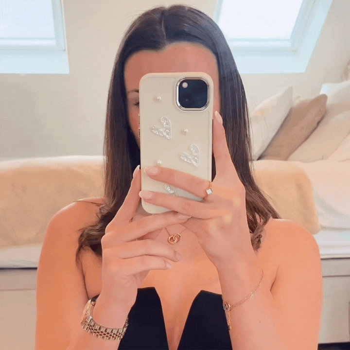 A person with long brown hair is taking a mirror selfie with their iPhone 15, which is adorned in Coconut Lane's Pearly Hearts Phone Case - Nude. They are wearing a strapless black top along with an assortment of jewelry including rings, bracelets, and a necklace. The background features a bed with pillows.