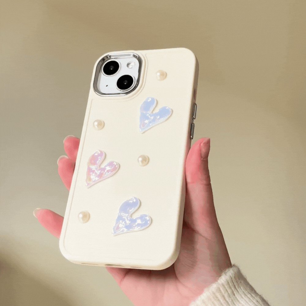 A hand holds the Pearly Hearts Phone Case - Nude from Coconut Lane, featuring an extra protective, cream-colored design adorned with pastel hearts and small pearl-like beads against a neutral background. The phone case boasts a textured design, and the person appears to be wearing a light-colored sweater.
