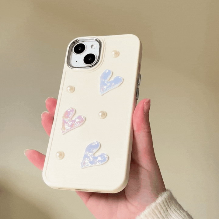 A hand holds the Pearly Hearts Phone Case - Nude from Coconut Lane, featuring an extra protective, cream-colored design adorned with pastel hearts and small pearl-like beads against a neutral background. The phone case boasts a textured design, and the person appears to be wearing a light-colored sweater.

