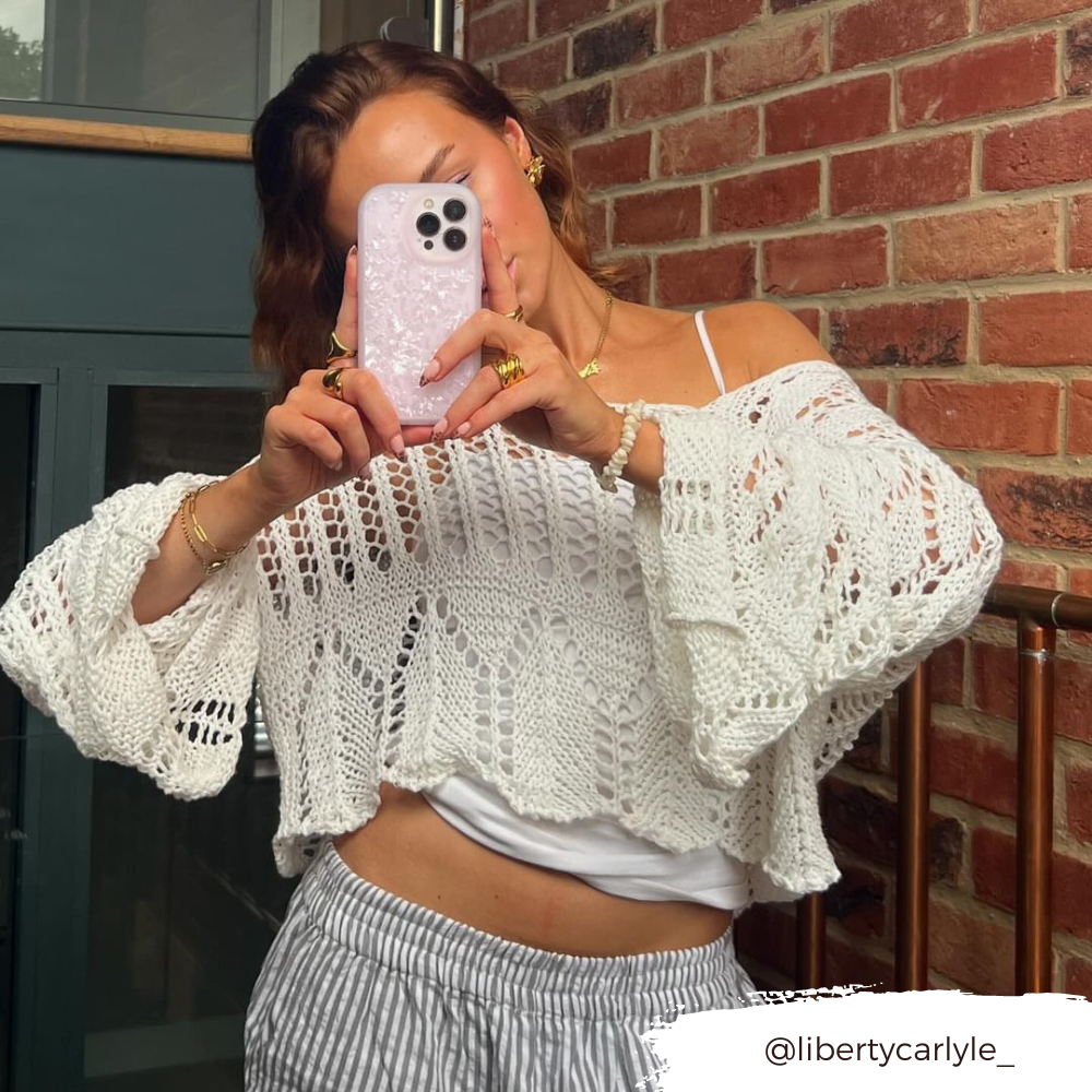 A person dressed in a loose white crochet sweater layered over a white top holds up an iPhone15 adorned with the Coconut Lane Moonstone Pink Phone Case, capturing a mirror selfie. They stand in front of a brick wall with part of a dark green door visible. The Instagram handle "@libertycarlyle_" is displayed at the bottom right.