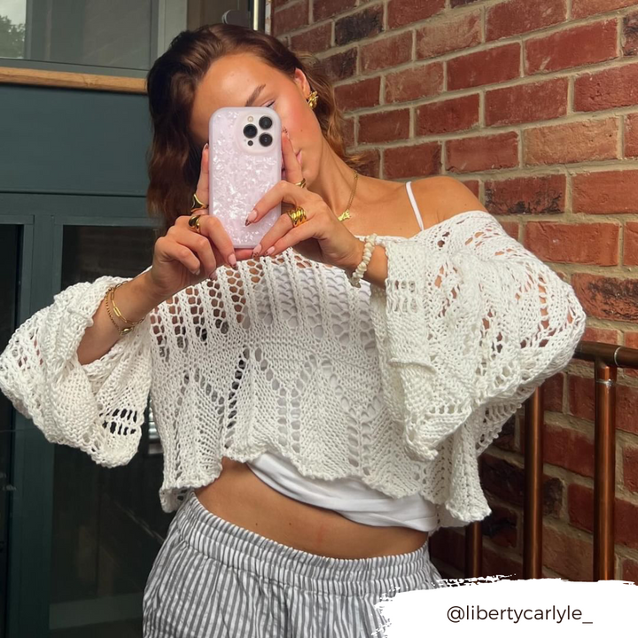 A person dressed in a loose white crochet sweater layered over a white top holds up an iPhone15 adorned with the Coconut Lane Moonstone Pink Phone Case, capturing a mirror selfie. They stand in front of a brick wall with part of a dark green door visible. The Instagram handle "@libertycarlyle_" is displayed at the bottom right.