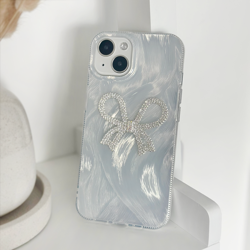 A close-up of a white iPhone with a dual-camera setup, encased in Coconut Lane's Diamante Bow Phone Case—a clear phone case adorned with a sparkly silver diamante bow design. The unique case not only adds flair but also provides extra protection. The phone is propped against a white surface, with a beige object partially visible in the background.