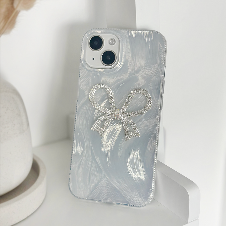 A close-up of a white iPhone with a dual-camera setup, encased in Coconut Lane's Diamante Bow Phone Case—a clear phone case adorned with a sparkly silver diamante bow design. The unique case not only adds flair but also provides extra protection. The phone is propped against a white surface, with a beige object partially visible in the background.