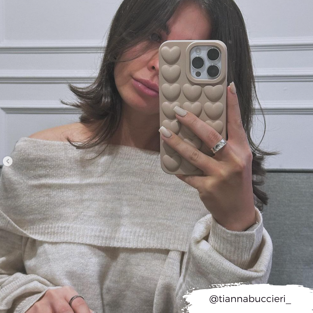 A woman takes a mirror selfie, holding an iPhone15 encased in the Coconut Lane 3D Hearts Phone Case - Matte Nude. She wears an off-shoulder beige sweater and has her nails painted in a neutral color. The corner of the photo features text that says "@tiannabuccieri_".