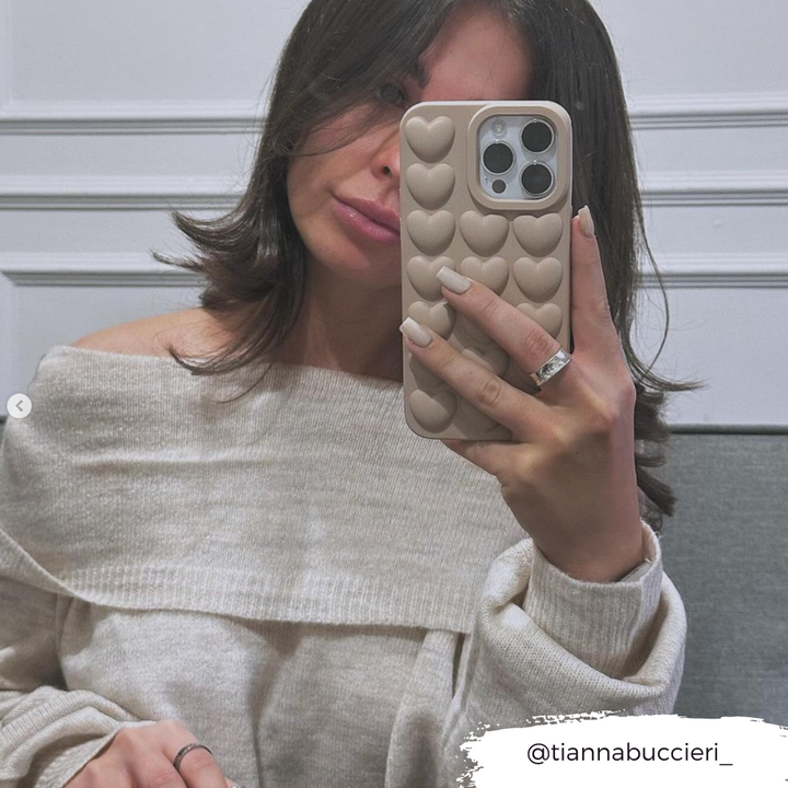 A woman takes a mirror selfie, holding an iPhone15 encased in the Coconut Lane 3D Hearts Phone Case - Matte Nude. She wears an off-shoulder beige sweater and has her nails painted in a neutral color. The corner of the photo features text that says "@tiannabuccieri_".