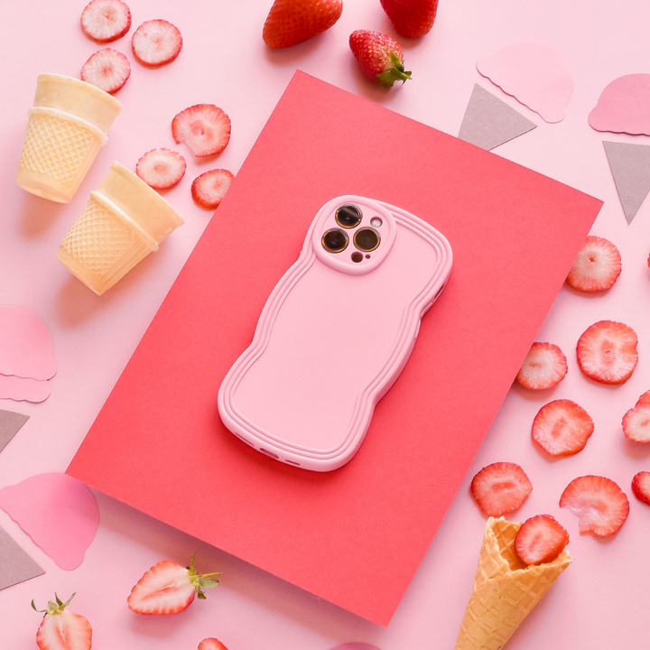 A pink smartphone encased in a Coconut Lane NAKD Curvy Phone Case - Strawberry, showcasing a textured, matte finish, is placed on a red sheet against a pink background. Surrounding it are sliced strawberries, whole strawberries, ice cream cones, and cutouts of ice cream shapes, creating a colorful dessert-themed arrangement.