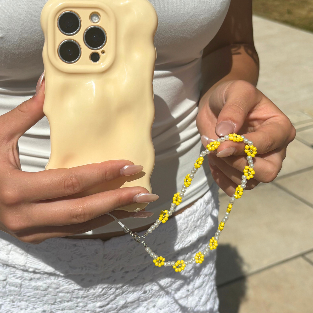 Beige wavy phone case with a sunflower-patterned beaded strap, held outdoors in sunlight.