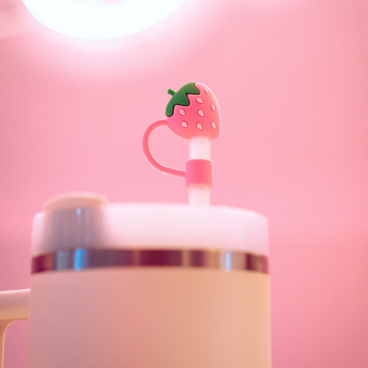 A white tumbler is adorned with a pink straw featuring Coconut Lane's Strawberry Straw Cover, a charming silicone topper shaped like a strawberry. The background is softly illuminated in pink hues by a circular light fixture, creating a cozy and playful atmosphere.