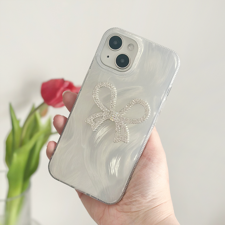 A hand holding a smartphone featuring the Diamante Bow Phone Case by Coconut Lane, an extra protective, pearlized white case adorned with a sparkly diamante bow design. In the blurred background, there's a red tulip in a clear vase.