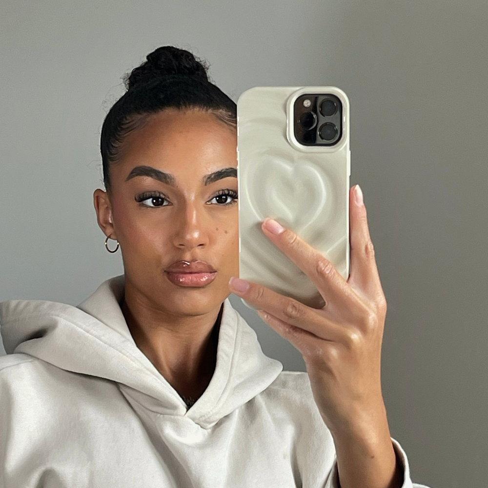 A person with neatly styled hair in a bun takes a selfie using a smartphone adorned with Coconut Lane's Melting Heart Phone Case in Coconut Cream. They are wearing a light-colored hoodie and gold hoop earrings, maintaining a neutral facial expression against a plain background.
