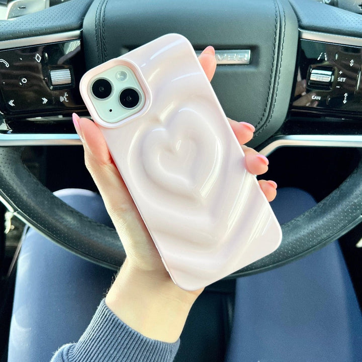 A person with pink nail polish holds a smartphone adorned with Coconut Lane's Melting Heart Phone Case in Peony Pink. Seated in the driver's seat of a vehicle, the steering wheel and dashboard are visible in the background, highlighting the chic allure of their phone case featuring a raised heart design.
