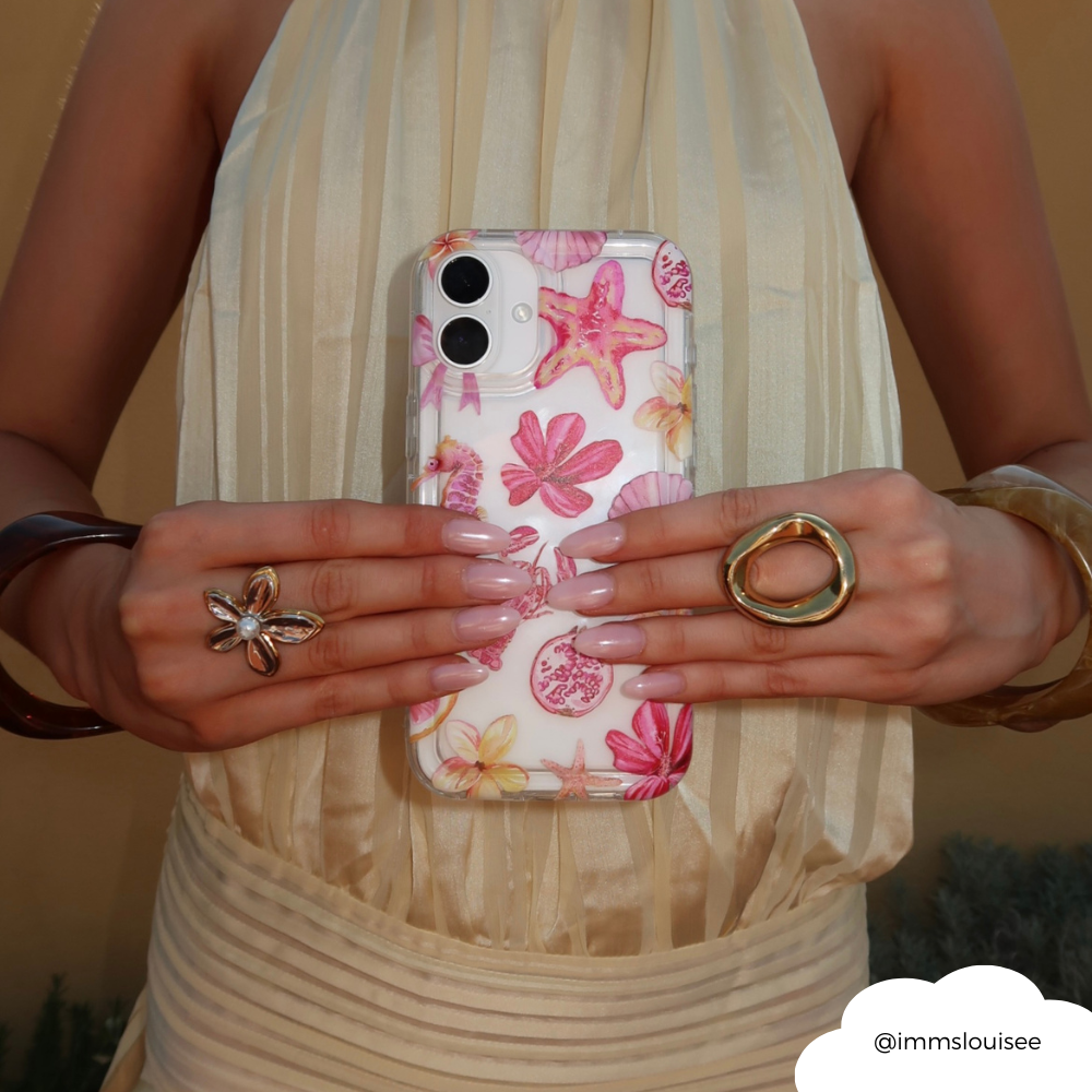 Hand holding phone in Coconut Lane clear case with Rosé Coast design; cream dress, manicured nails, bold rings visible.
