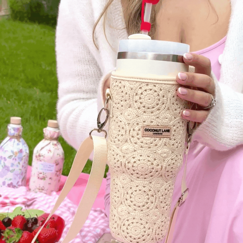 A person dressed in a light pink outfit holds a large beverage in the chic Coconut Lane Tumbler Carry Case - Festival Crochet, featuring an elegant strap. In the background, there's a picnic scene with a pink checkered blanket, fresh strawberries, and two floral-patterned bottles. Grass and trees complete the picturesque setting.