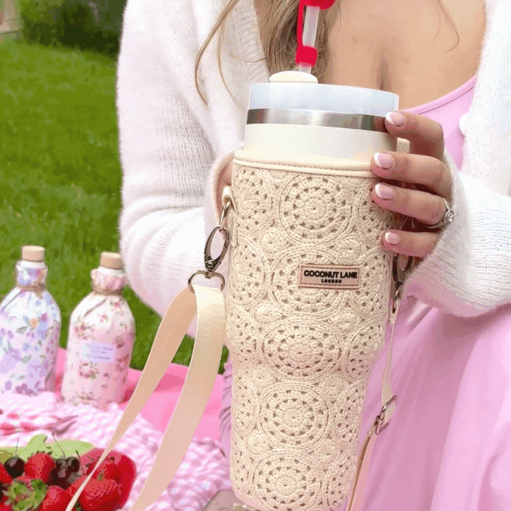 A person dressed in a light pink outfit holds a large beverage in the chic Coconut Lane Tumbler Carry Case - Festival Crochet, featuring an elegant strap. In the background, there's a picnic scene with a pink checkered blanket, fresh strawberries, and two floral-patterned bottles. Grass and trees complete the picturesque setting.