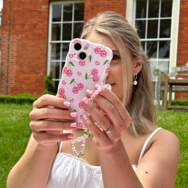 A person with long, light-colored hair is holding a smartphone encased in Coconut Lane's Wavy Phone Case - Disco Cherries, complete with an attached beaded bracelet. She is taking a photo outdoors in front of a building with large windows and a grassy lawn, showcasing her pearl earring and manicured nails.