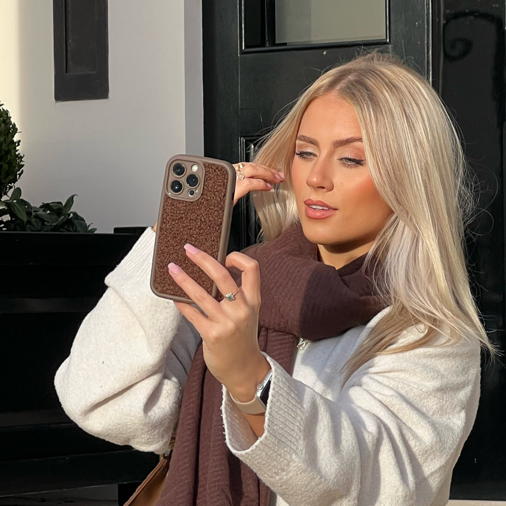 A person with long, blonde hair is taking a selfie with their smartphone, encased in the extra protective Cosy Teddy Phone Case - Brown by Coconut Lane. They are wearing a white coat and a brown scarf, standing outside in front of a black door. The lighting highlights their features and hair beautifully.