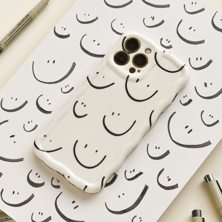 A Coconut Lane Wavy Phone Case - Smiley Doodle in white, adorned with simple black smiley faces, rests on a sheet of white paper covered with analogous smiley doodle designs. Various markers are scattered around the phone, merging seamlessly into the cheerful, creative backdrop.