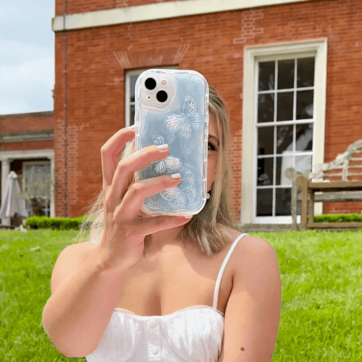 A woman in a white sleeveless dress takes a selfie with a phone encased in the Coconut Lane Shimmer Blue Butterflies Phone Case. She is outdoors in front of a brick building with large windows and green grass.