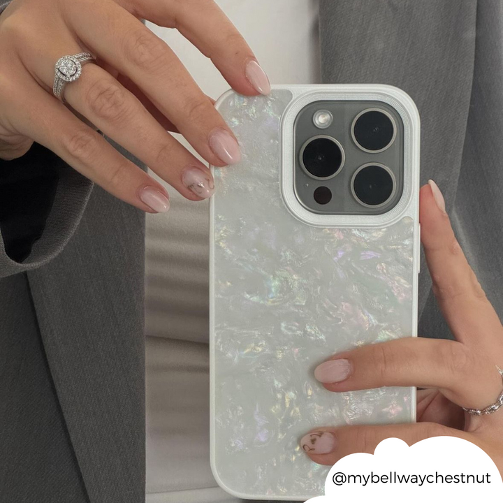 A person with a grey blazer is holding a Mother of Pearl Phone Case by Coconut Lane in their left hand. Their nails are painted in a light, natural color with minimalist designs. There is a diamond ring on their left ring finger, and the username "@mybellwaychestnut" is visible.
