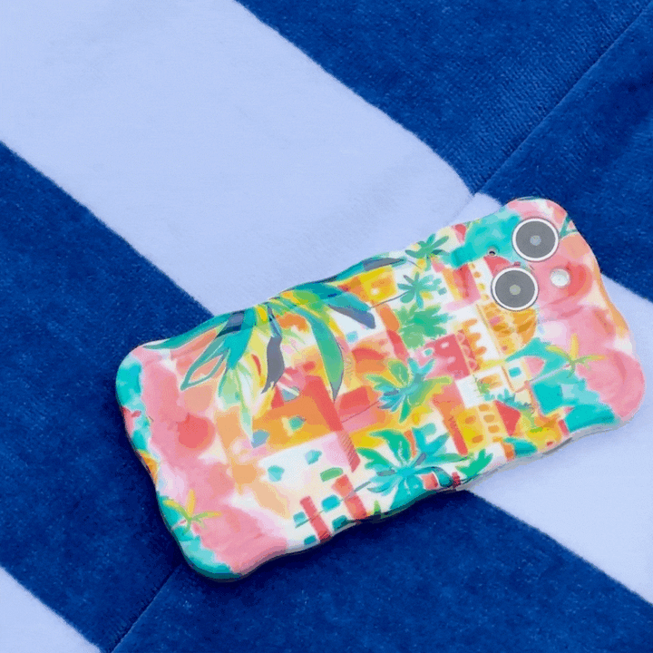 A Wavy Phone Case – Portofino Dreams by Coconut Lane, showcasing a colorful and vibrant Italian seaside design with palm trees and houses, rests on a surface adorned with blue and white stripes. The extra protective case includes cutouts for dual cameras and a flash.