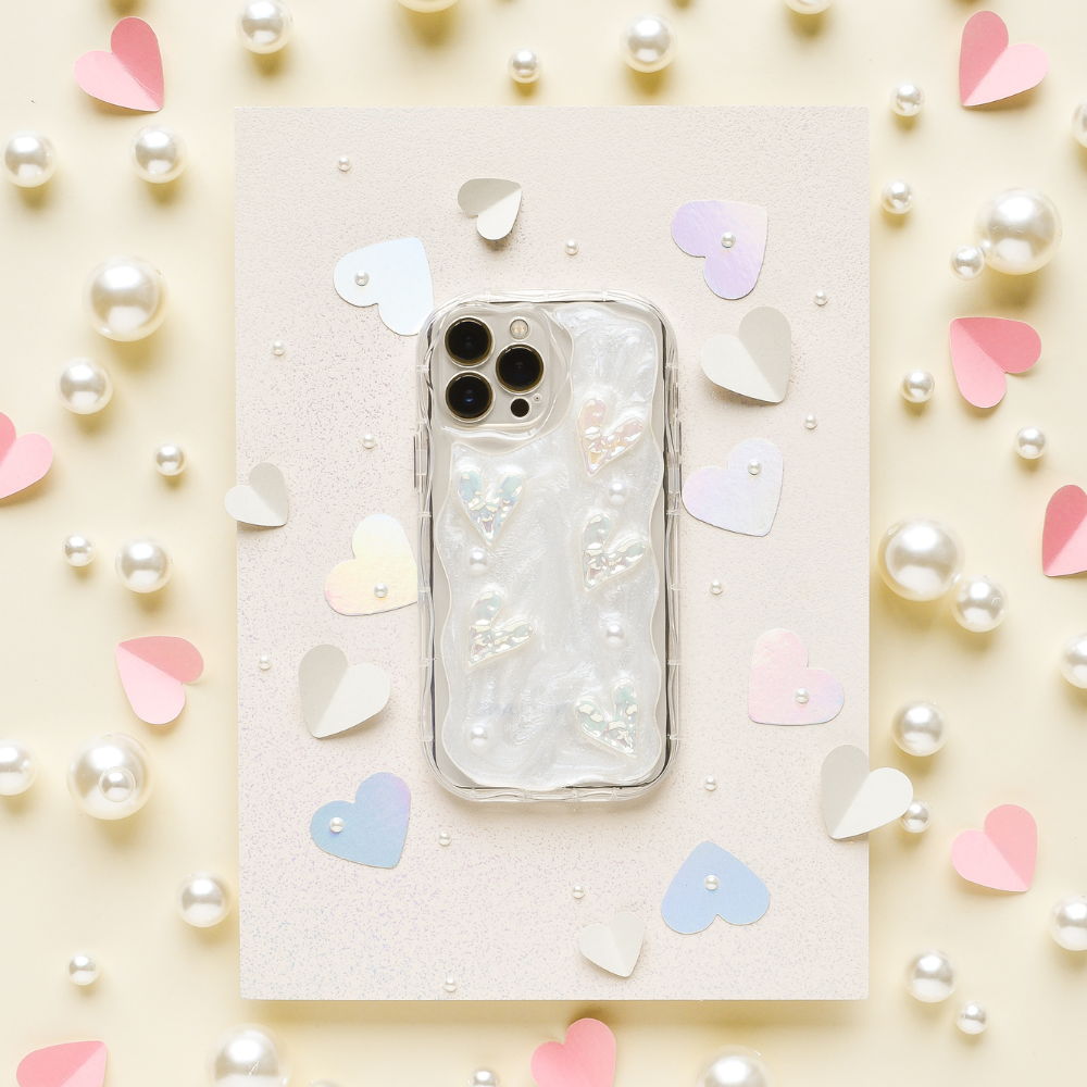 An iPhone15 with a Coconut Lane's Pearly Hearts Phone Case - Clear lies on a sparkly white surface. The background features scattered white pearls and cut-out heart shapes in pastel colors, including pink, lilac, and beige.