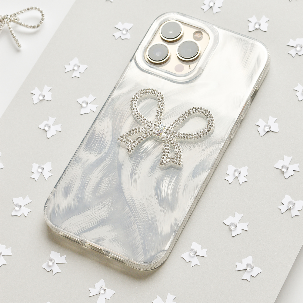 A smartphone encased in the Coconut Lane Diamante Bow Phone Case, which boasts a sparkling bow decoration, rests on a light gray surface. This distinctive phone case infuses an element of elegance, while the background is sprinkled with scattered white, star-like confetti. The phone's camera lenses are clearly visible, prominently situated at the top left corner.