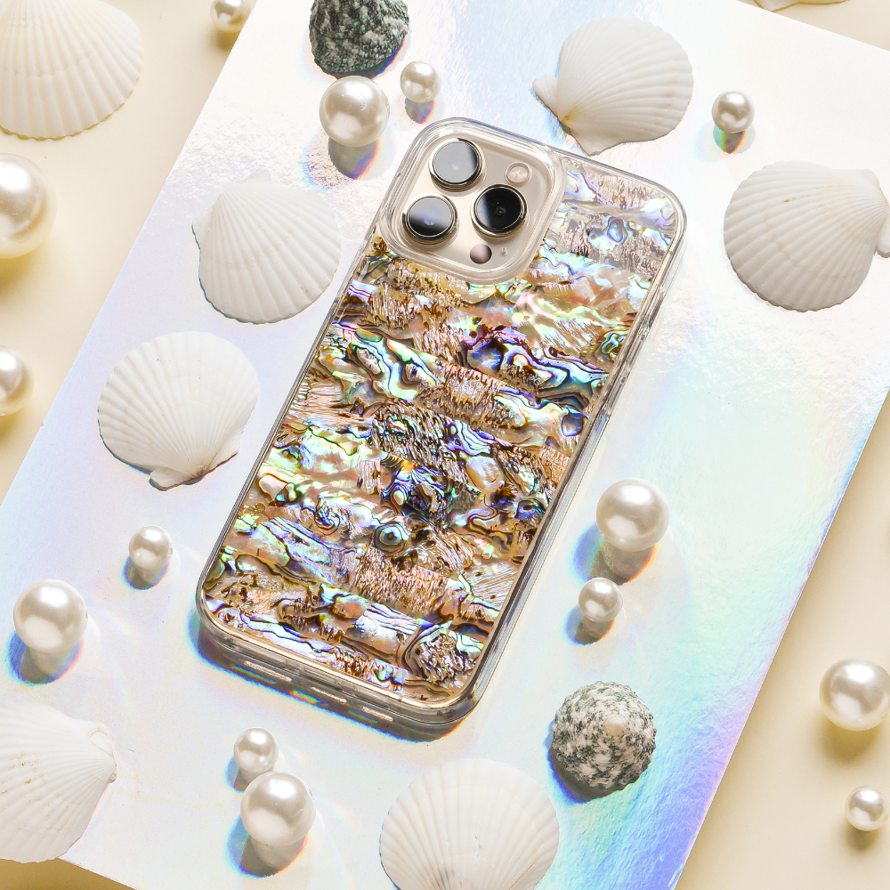 The Coconut Lane Real Natural Shell Phone Case - Black, showcasing an exquisite abalone shell-patterned design, is elegantly displayed on a reflective holographic surface. Surrounded by white seashells, spherical pearls, and textured grey stones, it crafts an immersive marine-inspired aesthetic.