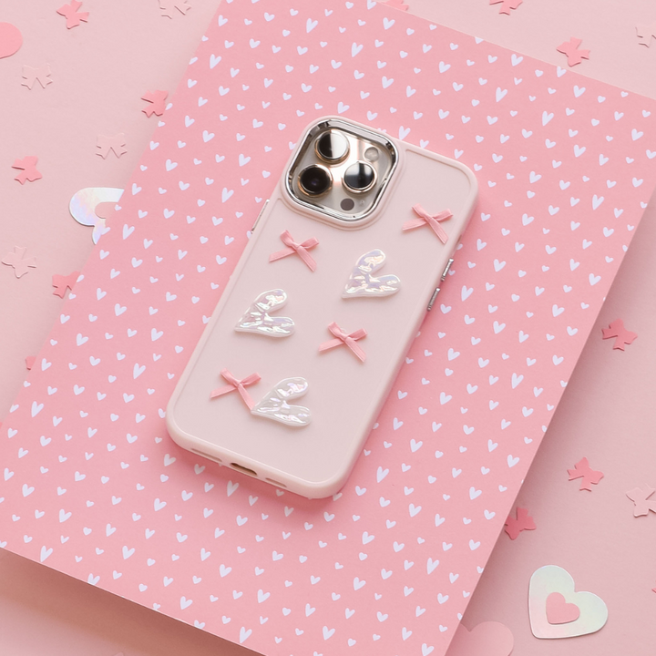 A Coconut Lane Pearly Hearts Phone Case - Pink, designed for the iPhone 15 and adorned with four white hearts and three pink bows, lies flat against a pink background featuring white heart patterns and scattered heart-shaped confetti. The phone's rear cameras are visible through cutouts in the case.