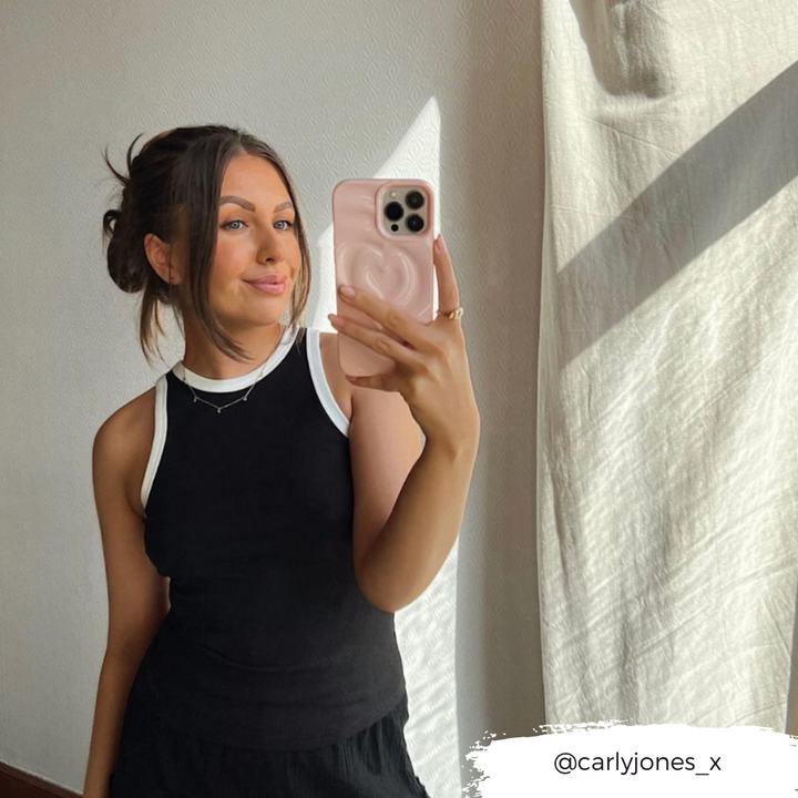 A woman taking a mirror selfie with her smartphone, adorned in the Coconut Lane Melting Heart Phone Case - Peony Pink. She is smiling with her hair styled in a bun with loose strands and is wearing a sleeveless black top with white trim. Her Instagram handle, @carlyjones_x, appears in the bottom right corner.