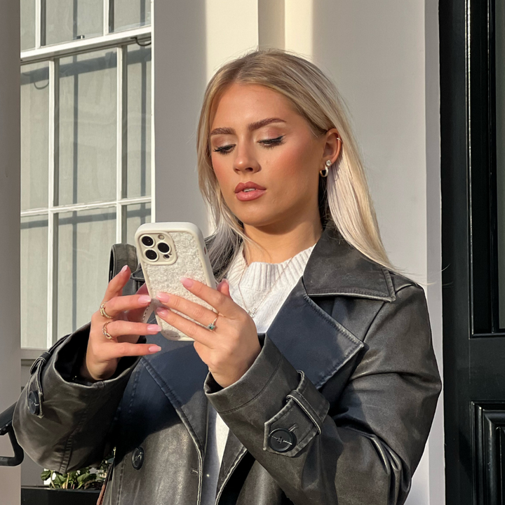 A woman with long, blonde hair stands outside a building, focused on her smartphone. She is wearing a dark leather jacket over a white top. Her Cosy Teddy Phone Case - Cream from Coconut Lane, featuring an extra protective, textured teddy finish, adds a unique touch. The black door and window with white frames in the background complement her attire.
