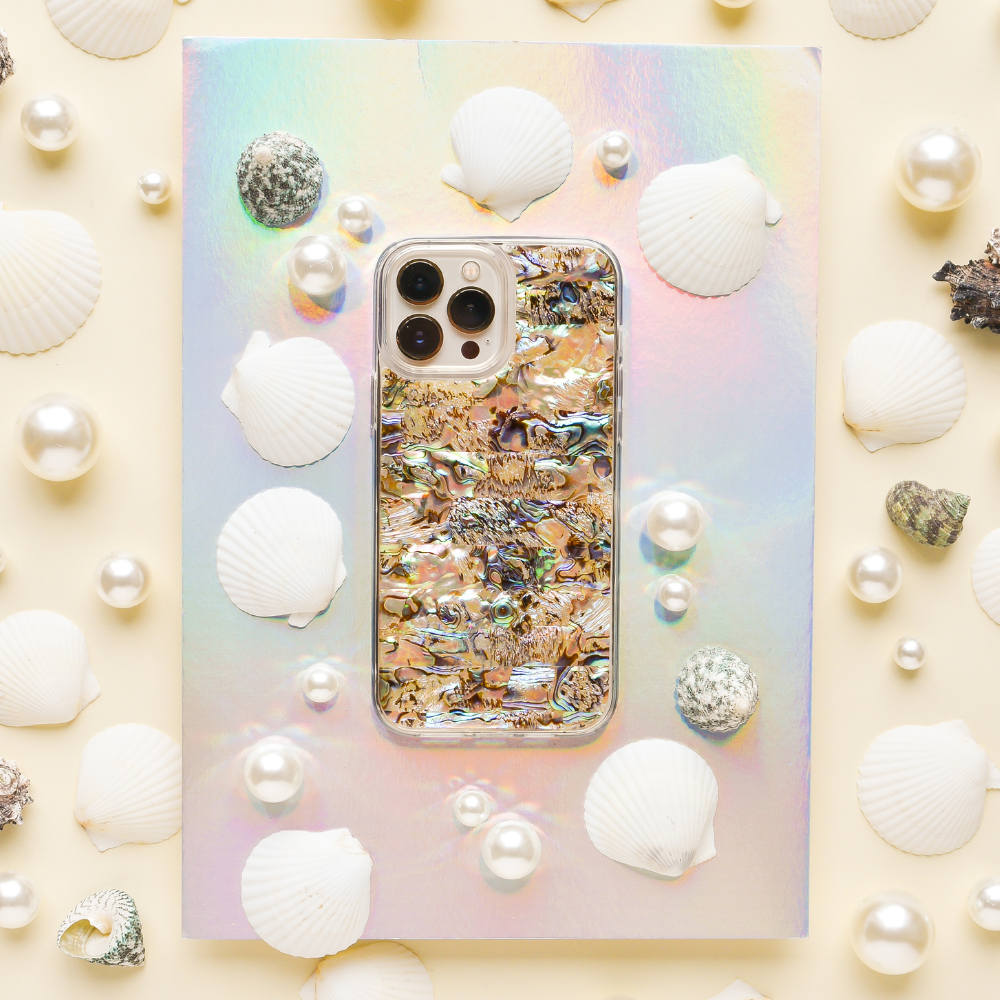 A smartphone with a colorful, iridescent case featuring an abstract design sits on a holographic surface. The phone, adorned with a Real Natural Shell Phone Case - Black from Coconut Lane, is surrounded by white seashells, pearls, and small textured stones, creating an ocean-inspired aesthetic.