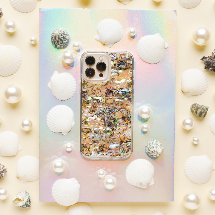 A smartphone with a colorful, iridescent case featuring an abstract design sits on a holographic surface. The phone, adorned with a Real Natural Shell Phone Case - Black from Coconut Lane, is surrounded by white seashells, pearls, and small textured stones, creating an ocean-inspired aesthetic.