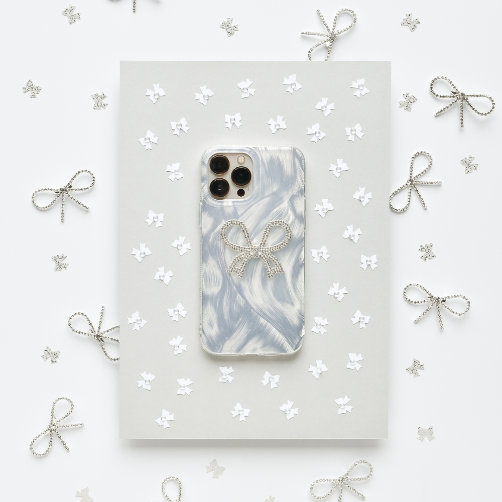 A Coconut Lane Diamante Bow Phone Case, featuring a marbled white and gray design with an extra protective decorative bow, is placed on a gray sheet surrounded by small paper stars and silver wire bows. The white background adds to the clean and elegant aesthetic.