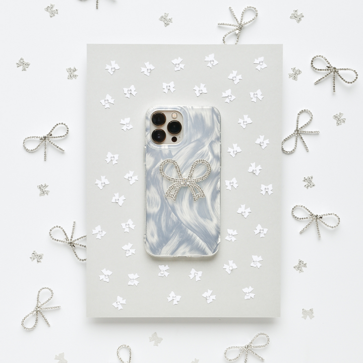 A Coconut Lane Diamante Bow Phone Case, featuring a marbled white and gray design with an extra protective decorative bow, is placed on a gray sheet surrounded by small paper stars and silver wire bows. The white background adds to the clean and elegant aesthetic.