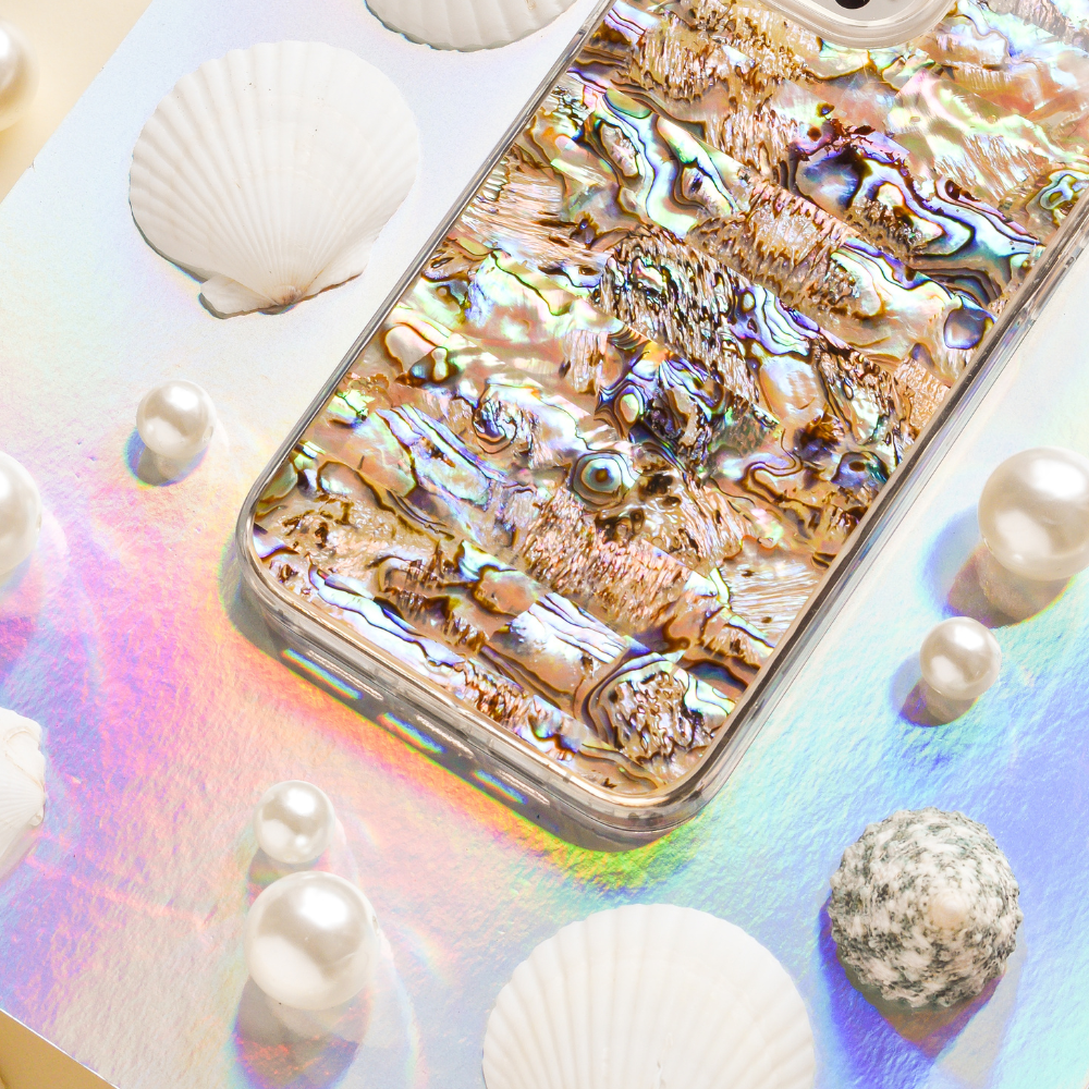 A smartphone with a reflective Coconut Lane Real Natural Shell Phone Case in Black lies on a pastel-colored, iridescent surface. Surrounding the phone are authentic durable shells and variously sized pearls, enhancing the aquatic theme.