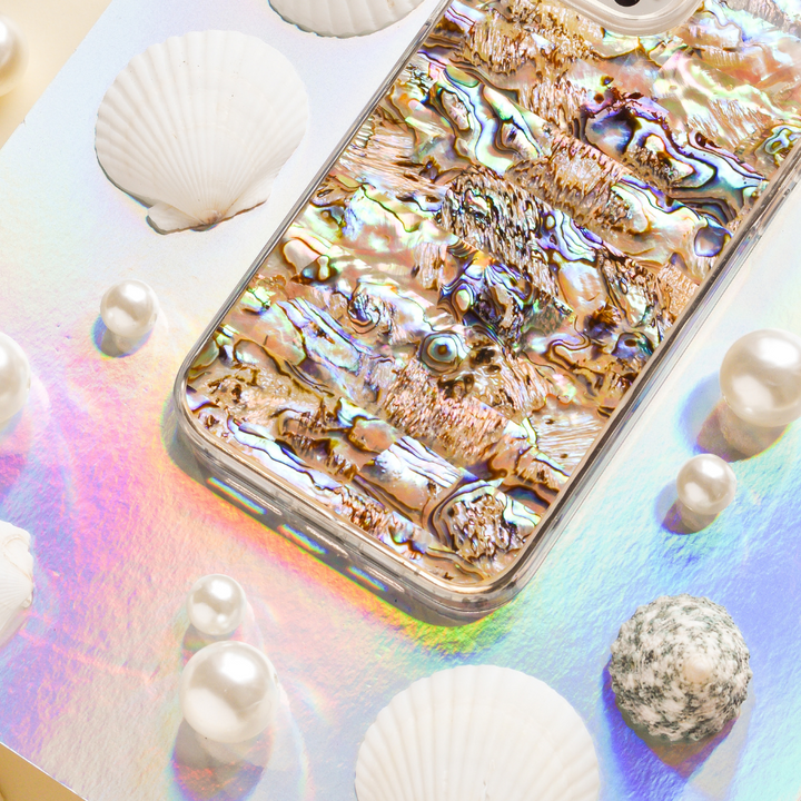 A smartphone with a reflective Coconut Lane Real Natural Shell Phone Case in Black lies on a pastel-colored, iridescent surface. Surrounding the phone are authentic durable shells and variously sized pearls, enhancing the aquatic theme.