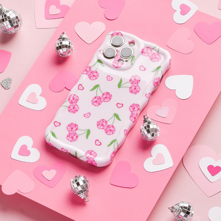 A Wavy Phone Case - Disco Cherries from Coconut Lane lies on a pink surface adorned with various paper hearts and small silvery disco balls. The case, featuring a playful cherry pattern on a white background, exudes a whimsical, romantic vibe.