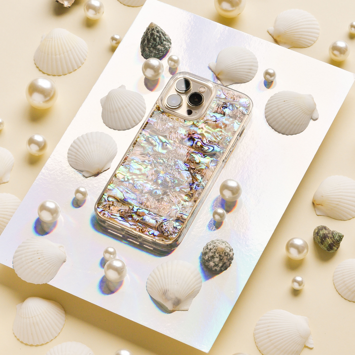 A shiny, holographic phone case named "Real Natural Shell Phone Case - Black" by Coconut Lane is displayed on a reflective surface, surrounded by white seashells and scattered pearls. The background is a light beige, creating a seaside-inspired aesthetic. The phone's camera lenses are visible through the genuine natural shell case.