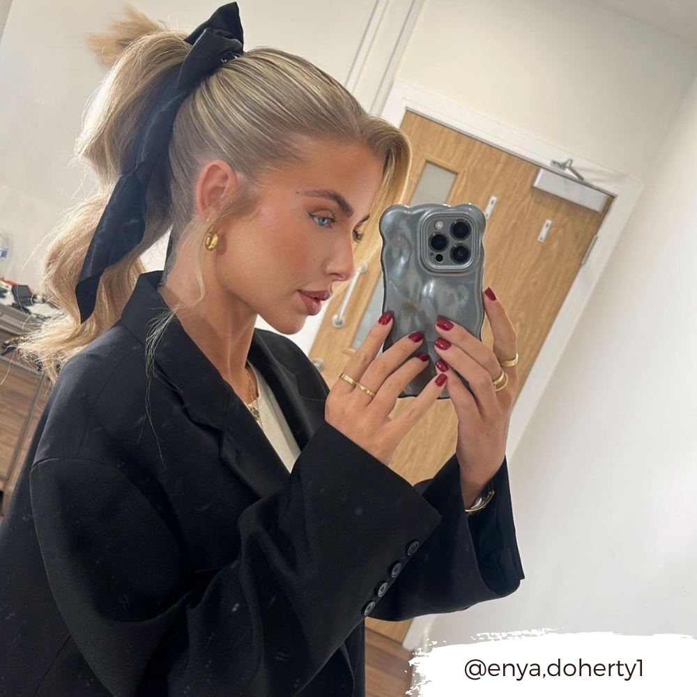 A woman with blonde hair tied in a ponytail with a large black bow takes a mirror selfie. She is wearing a black blazer over a white top and has red nail polish. She is holding an extra protective 3D Pearl Phone Case - Grey from Coconut Lane with a bear-shaped design. The handle @enya.dohertyl is written in the corner.