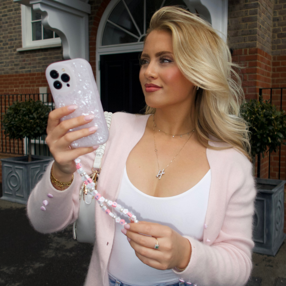 A woman with blonde hair, dressed in a pink cardigan and white top, holds up her smartphone encased in a Coconut Lane Moonstone Pink Phone Case to take a selfie. She stands in front of a brick building with arched windows and decorative plants, smiling slightly as she looks at the phone's screen.