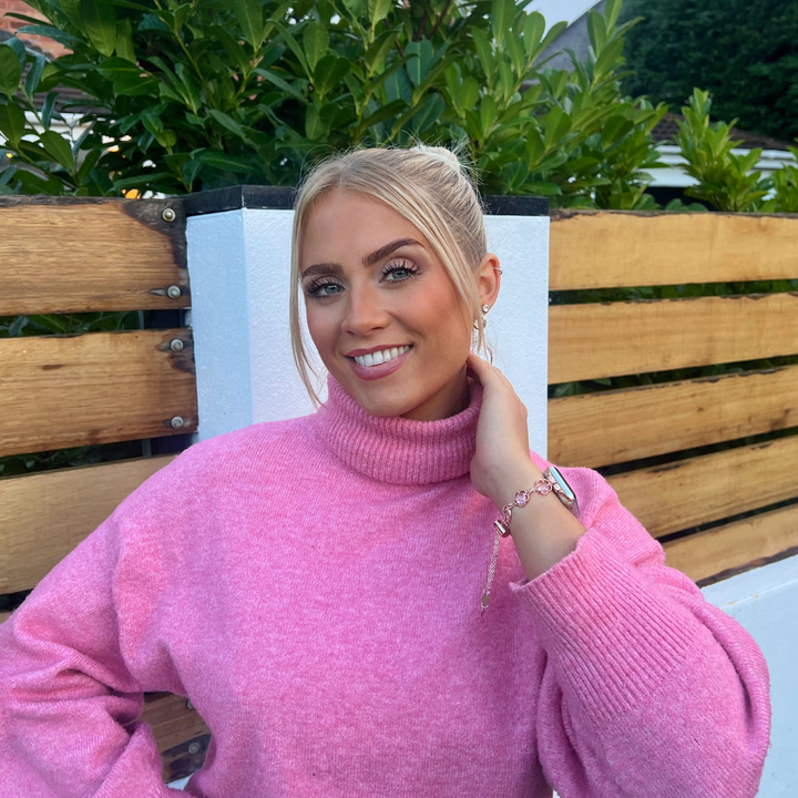 A blonde woman smiles as she stands in front of a wooden fence adorned with green foliage. She is dressed in a light pink turtleneck sweater and touches her neck, highlighting the Coconut Lane Chain Bracelet Apple Watch Strap - Rose Gold that perfectly complements her look. In the background, elements of a white wall and building are visible.